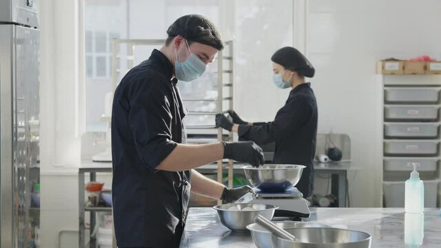 Side view of man in coronavirus face mask beating eggs for pastry in slow motion as blurred woman passing with commercial tray at background. Concentrated cook preparing dessert in kitchen.