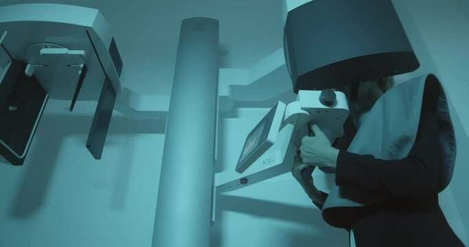 A female patient having 3D scanning of her jaw, she is standing inside of CBCT which is rotating around of her head and her mouth is open, her hands are in the special holder. 