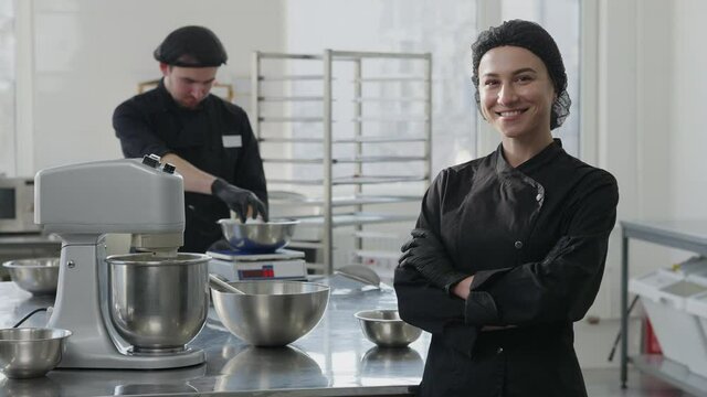 Portrait of charming confident young woman in cook uniform crossing hands looking at camera smiling with automatic mixer working at background and man weighing eggs for pastry dough. Culinary concept.