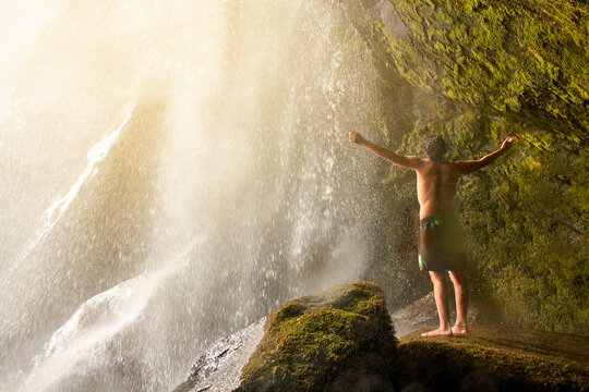Young man standing in a cave inside a waterfall. Man getting wet with arms open. Freedom and travel concept. © nicuervo