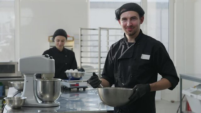 Middle shot portrait of young Caucasian man posing with bowl and whisk as blurred woman weighing eggs at background. Positive smiling baker at workplace with dessert ingredients. Job and culinary.