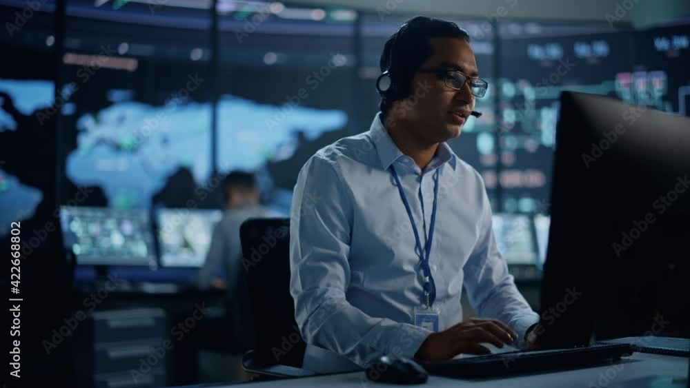 Poster Portrait of Professional IT Technical Support Specialist Working on Computer in Monitoring Control Room with Digital Screens. Employee Wears Headphones with Mic and Talking on a Call.