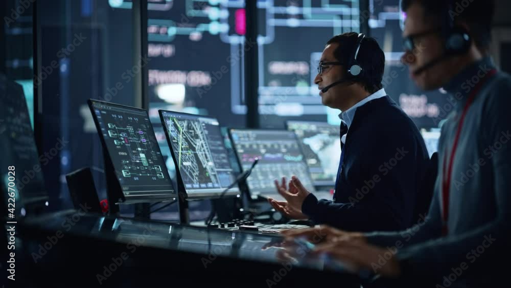Poster Portrait of Professional IT Technical Support Specialist Working on Computer in Monitoring Control Room with Digital Screens. Employee Wears Headphones with Mic and Talking on a Call.