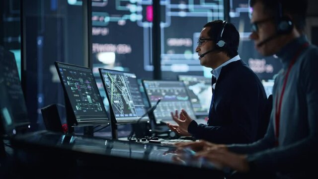 Portrait of Professional IT Technical Support Specialist Working on Computer in Monitoring Control Room with Digital Screens. Employee Wears Headphones with Mic and Talking on a Call.