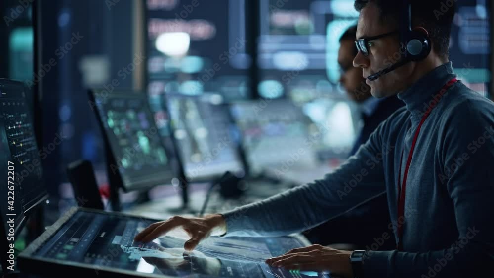 Poster Portrait of Professional IT Technical Support Specialist Working on Computer in Monitoring Control Room with Digital Screens. Employee Wears Headphones with Mic and Talking on a Call.