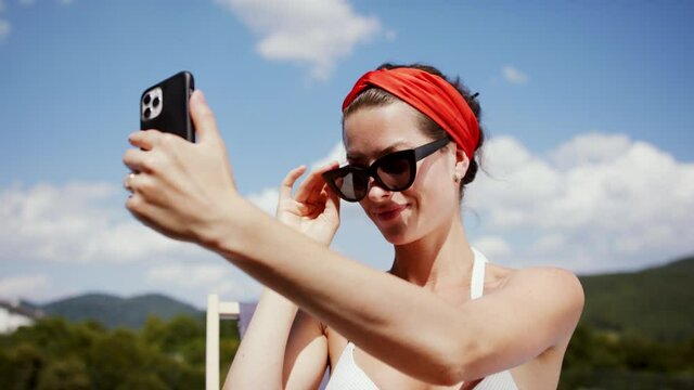 Young woman sitting by swimming pool outdoors in backyard garden, taking selfie.