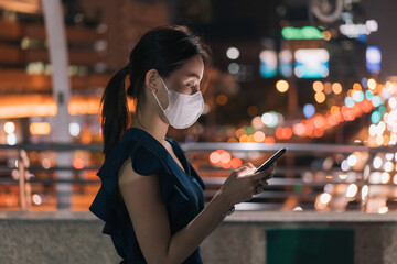 Side-view of beautiful young Asian happy woman wearing covid-19 face mask standing over footbridge checking smartphone and browsing through apps while checking email