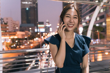 Portrait of smiling young Asian successful woman in casual clothing talking over smartphone while standing on footbridge with city view in background