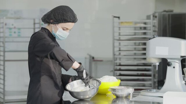 Side view of confident beautiful woman in coronavirus face mask mixing confectionery topping in slow motion. Charming Caucasian female baker working in commercial kitchen on Covid-19 pandemic.
