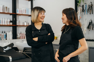 Happy young Asian female hairstylist colleagues working in salon standing wearing apron and...