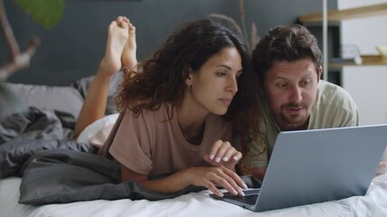 Young Caucasian couple lying together on bed at home, surfing the Internet and discussing something on laptop screen - Powered by Adobe