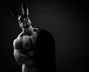 Naked topless muscular man in underwear, black rabbit mask and with jacket on shoulder stands at copy space over dark background - 422679627