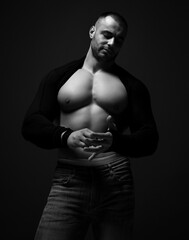 Fototapeta na wymiar Muscular man, athlete, bodybuilder in jeans and shirt with longsleeves stands with naked chest, looking down on dark background. Black and white portrait