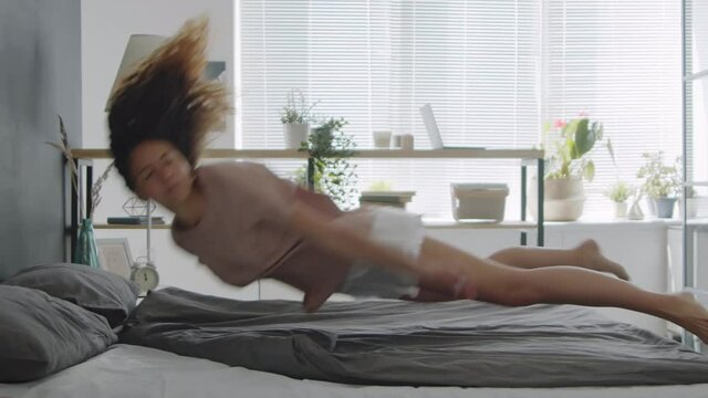 Slow motion shot of young barefoot woman in pajamas jumping on bed with eyes closed and sleeping on her stomach