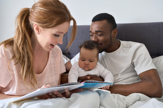 Happy young caucasian mom holding book reading stories to cute small mixed race child daughter in bed. Multiethnic family parents and their baby playing, having fun waking up in the morning together.