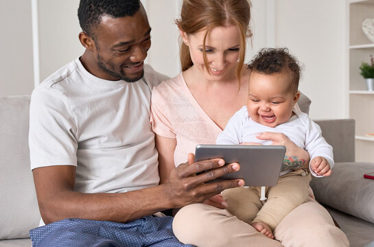 Happy young multiethnic family couple holding cute infant daughter using digital tablet sitting on couch. Funny mixed race child baby girl laughing watching cartoons with diverse parents at home.