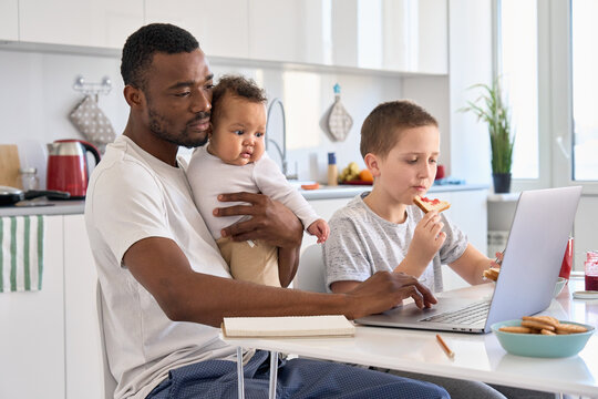 Young Black father, African dad remote working or distance learning online from home office using laptop computer having breakfast holding mixed race baby sitting at kitchen table with kid son at home