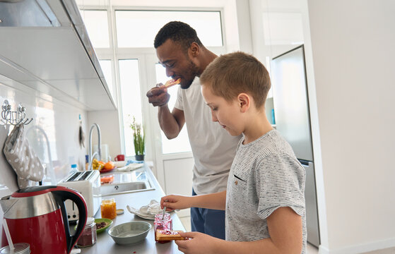 Hungry teenage Caucasian boy son helping young African American Black father cooking together making and eating toasts with jam on mixed race family breakfast or snack standing in kitchen at home.