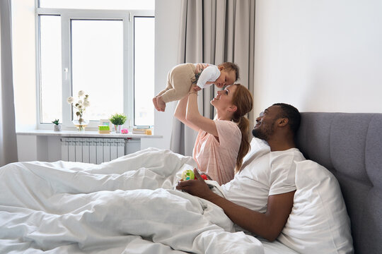 Multiracial diverse young family couple playing with cute baby child in bed holding infant kid girl. Happy multiethnic parents having fun in the morning lifting small daughter in bedroom at home.