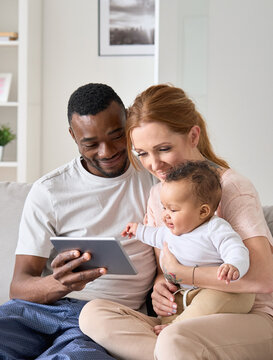 Happy interracial diverse parents holding small cute mixed race baby kid daughter learning using digital tablet computer sitting on sofa at home spending time with technology device at home together.