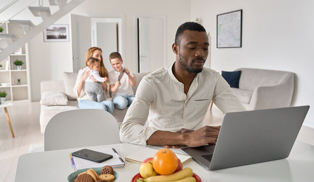 Young Black parent dad distance working from home office with family. African American father using laptop computer spending time with diverse multiracial kids and wife in modern house living room.