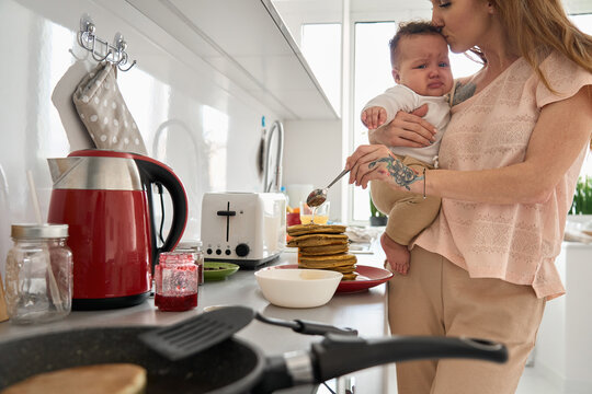 Young mother preparing pancakes pouring with honey holding cute little baby soothing crying child daughter standing in kitchen at home. Mom and infant kid cooking homemade family breakfast together.