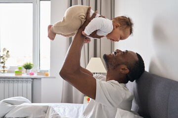 Happy affectionate young Black dad lifting adorable mixed race baby daughter having fun in bed at...