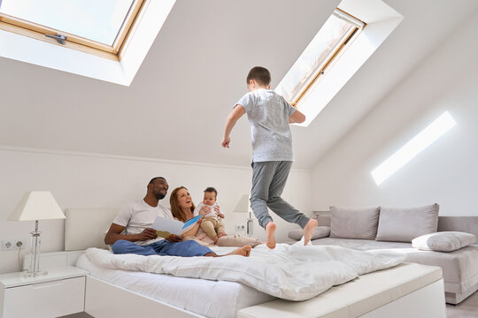 Happy multiracial family with kids having fun in sunny bedroom waking up together. Smiling interracial mom and dad reading book to baby daughter laughing while teen son jumping on bed playing at home.
