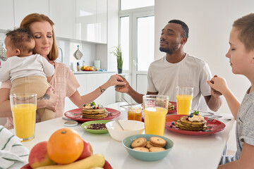 Happy multiracial family couple with children pray together before having morning breakfast at home...