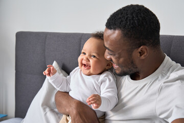 Happy loving young Black dad holding adorable mixed race baby daughter having fun in bed at home....
