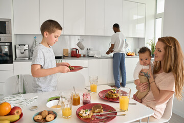 Happy multiracial diverse family couple with kids having breakfast in kitchen. Caucasian teen boy son helping African father clearing table and washing dishes while mom and baby eating meal at home.