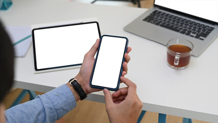 Close up view of businessman using multiple devices at workplace and holding smart phone with blank screen.