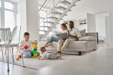 Multiethnic family spending time together in new home. Mixed race diverse children playing toys on carpet while multiracial parents using laptop and reading book sitting on couch in house living room.