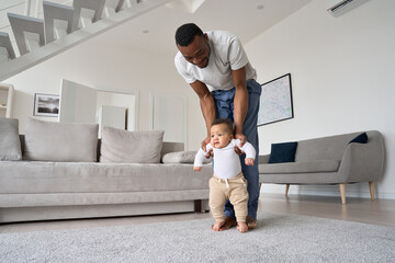 Young Black father helping cute infant child daughter teaching kid to walk at home. Mixed race baby learning doing first steps with loving African American dad playing together in living room. - Powered by Adobe