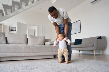 Young Black father helping cute infant child daughter teaching kid to walk at home. Mixed race baby learning doing first steps with loving African American dad playing together in living room.