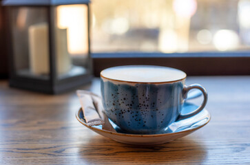 blue cup with coffee and foam in the form of a heart on a table with a bag of sugar on the background of a window during the day