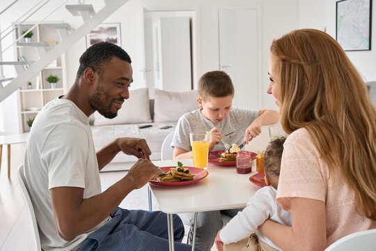 Happy interracial diverse family couple with two children having morning breakfast at home together. Multiethnic young parents with mixed race kids enjoying eating pancakes sitting at kitchen table. - Powered by Adobe