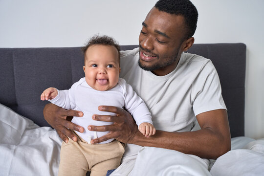 Happy Young Black Dad Holding Adorable Mixed Race Baby Daughter Having Fun In Bed At Home. Smiling African American Father Playing With Cute Funny Infant Child Girl Waking Up In Bedroom In The Morning