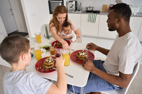 Happy Multiethnic Family With Kids Having Pancakes Meal On Breakfast Sitting At Kitchen Table In The Morning. Mixed Race Young Parents And Diverse Children Eating Cooked Food Together At Home.