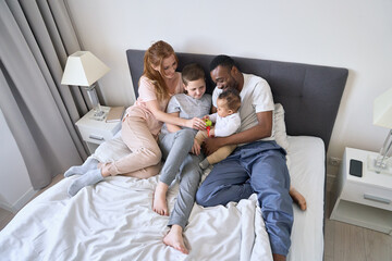 Happy multiethnic family with kids having fun waking up in bed together. Smiling parents African dad and Caucasian mom playing with mixed race diverse children in bedroom together at home, top view.