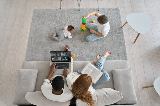 Multiethnic Family Spending Time Together At Home. Mixed Race Diverse Children Playing On Carpet While Parents Having Virtual Meeting Using Laptop And Reading Sitting On Couch. Top View From Above