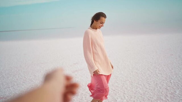 POV first person view beautiful happy woman in big kozy sweater and pink dress walking at windy salty coastline on pink salt lake. person gives hand to woman, follow me concept