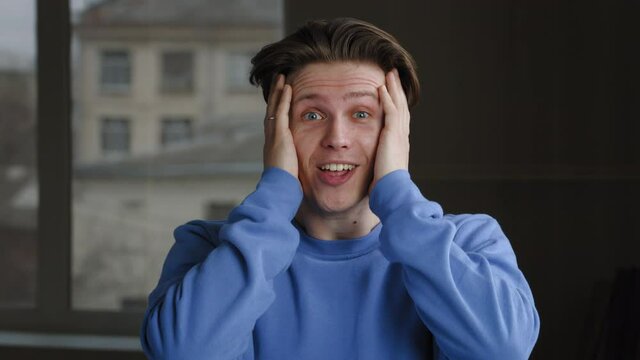 Close-up of male surprised face looking at camera, portrait of shocked amazed man caucasian guy millennial teenager wears blue sweatshirt says wow opens mouth holding his head from incredible news