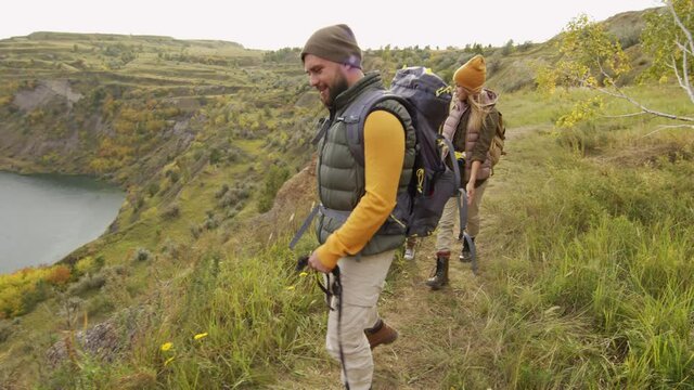 Tracking slowmo shot of happy family and cute dog on leash hiking near quarry lake and enjoying scenery. They are walking along narrow path on hill and chatting on windy day