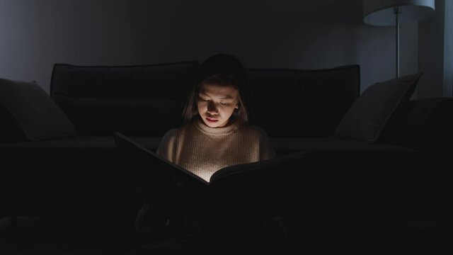 Night time, portrait of asian female sitting at home on the floor and reading a book, warm light, self-isolation during the pandemic, mystical lighting effect.