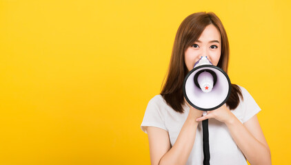 Fototapeta premium Asian happy portrait beautiful cute young woman teen standing making announcement message shouting screaming in megaphone looking to camera isolated, studio shot on yellow background with copy space