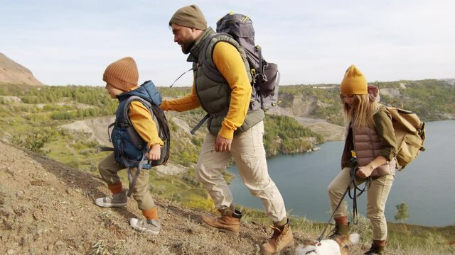 Slowmo tracking of bearded man with backpack holding hand of cute 5-year-old boy and helping him walk uphill during hike in scenic location. Young woman with dog leash walking behind them
