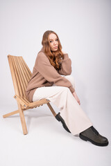 Fototapeta na wymiar Beautiful woman with curly red hair in beige pants and a taupe hoodie sitting in wooden armchair on studio background.