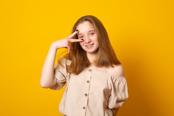 Contented smiling teen girl makes peace gesture over eye, shows two fingers, looks thoughtfully aside. Girl shows v sign.