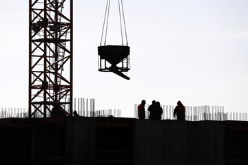 Silhouettes of workers and tower crane with cargo on construction site against the sky. Housing...